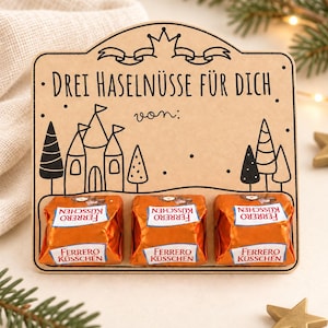 Op de afbeelding: Een rechthoekig lichtbruin houten displaybord met een zwarte lijntekening van een kasteel, bomen en een kroon. De tekst luidt "DREI HASELNÜSSE FÜR DICH" en "von:". Drie oranje verpakte Ferrero Küsschen chocolaatjes zijn aan de basis bevestigd.