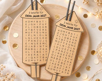 Könnte beinhalten: Zwei hellbraune, raketenförmige Pappkarten mit einem Buchstabengitter und dem Text 3 Worte für DEIN JAHR 2027. Jede Karte ist mit zwei dünnen grauen Wunderkerzen oben und unten versehen, die für Silvesterfeiern gedacht sind.