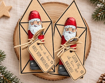 May include: Two house-shaped kraft paper cards featuring a chocolate Santa Claus wrapped in red foil. Each card is tied with jute twine and includes a small rectangular tag. The tags display the German text: das ist das HAUS VOM NIKOLAUS and GUTEN TAG, ich bin der NIKOLAUS!
