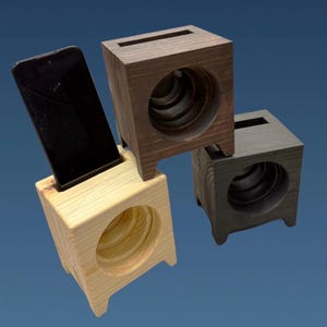 May include: Three cube-shaped wooden smartphone stands with a hollow circular centre and a top slot. One stand is light natural wood, one is medium brown, and one is dark charcoal. A black smartphone is inserted into the light wood stand.