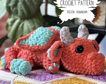 May include: Crochet pattern for a red and teal dragon amigurumi toy. The dragon is lying down with its wings spread out. The pattern is a digital download.