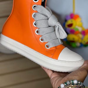 May include: High-top orange sneaker with a white toe cap and sole. The shoe features gray laces and silver eyelets. The sneaker is held in a hand, with a bracelet visible in the foreground. The shoe's design is modern and stylish.