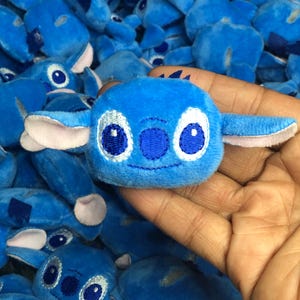 Op de afbeelding: Een close-up van een blauw pluche speelgoed met geborduurde ogen en een gestikte glimlach. Het speelgoed is gevormd als een cartoonfiguur met grote oren. Het speelgoed is omringd door andere vergelijkbare pluche speelgoed.