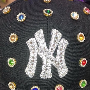 May include: Black baseball cap with a sparkling silver "NY" logo and colorful jewel accents. The cap features a curved brim and is adorned with various colored gems around the crown, adding a touch of glamour.