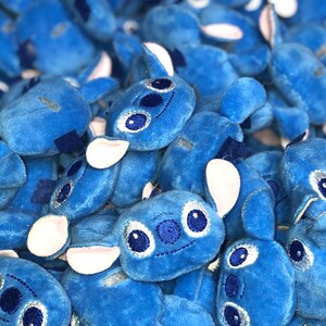 Op de afbeelding: Een stapel blauwe pluche Stitch-speelgoed met witte oren en geborduurde ogen. De speelgoedjes zijn allemaal naar de camera gericht en hebben een vrolijke uitdrukking.