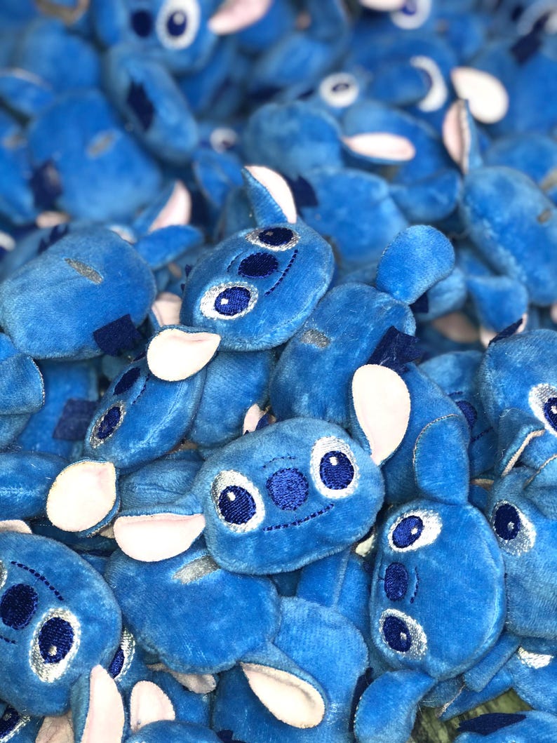 Op de afbeelding: Een stapel blauwe pluche Stitch-speelgoed met witte oren en geborduurde ogen. De speelgoedjes zijn allemaal naar de camera gericht en hebben een vrolijke uitdrukking.