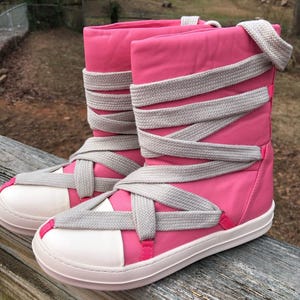May include: A pair of pink and white high-top boots with gray crisscrossing laces. The boots have a white sole and a pink upper, with gray laces wrapping around the shaft. The boots are made of a soft, padded material.