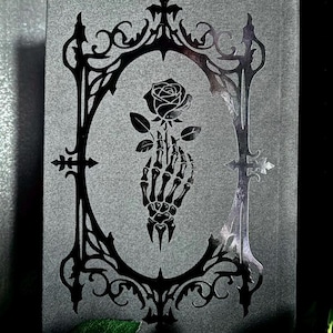 May include: A rectangular dark gray book cover featuring a glossy black foil-stamped design. The central graphic depicts a skeletal hand holding a single rose, enclosed within an ornate, pointed gothic-style frame with intricate filigree details.