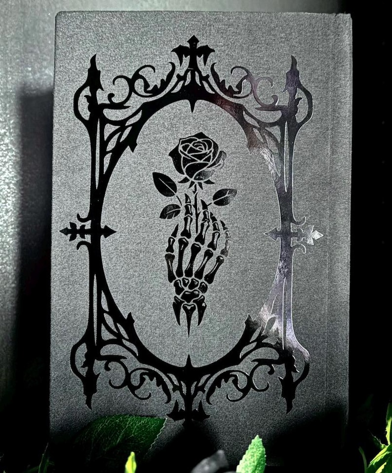 May include: A rectangular dark gray book cover featuring a glossy black foil-stamped design. The central graphic depicts a skeletal hand holding a single rose, enclosed within an ornate, pointed gothic-style frame with intricate filigree details.