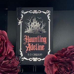 May include: A black hardcover book titled Haunting Adeline by H. D. Carlton, featuring a silver foil illustration of a house and trees, red lettering, and a black lace bookmark with a small silver charm attached to the top corner of the cover.