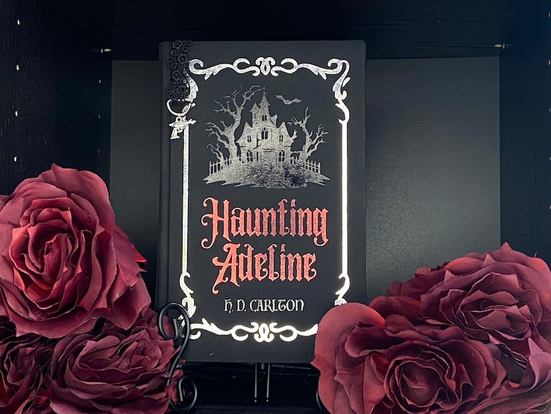 May include: A black hardcover book titled Haunting Adeline by H. D. Carlton, featuring a silver foil illustration of a house and trees, red lettering, and a black lace bookmark with a small silver charm attached to the top corner of the cover.