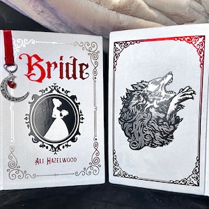 Puede incluir: Dos libros blancos de tapa dura con detalles plateados y rojos. El libro de la izquierda tiene la palabra "Bride" en letras rojas y el nombre del autor "Ali Hazelwood" en letras negras. El libro de la derecha tiene una ilustración de lobo plateado.