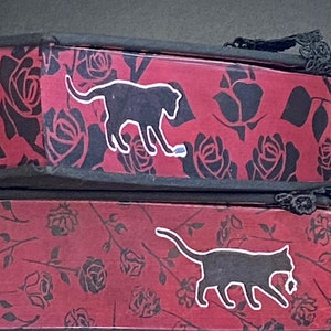 May include: Two stacked rectangular boxes featuring a red background with a black rose pattern. Each box displays a black cat silhouette with a white outline. The top box shows a cat playing with a small object, while the bottom box shows a cat walking.