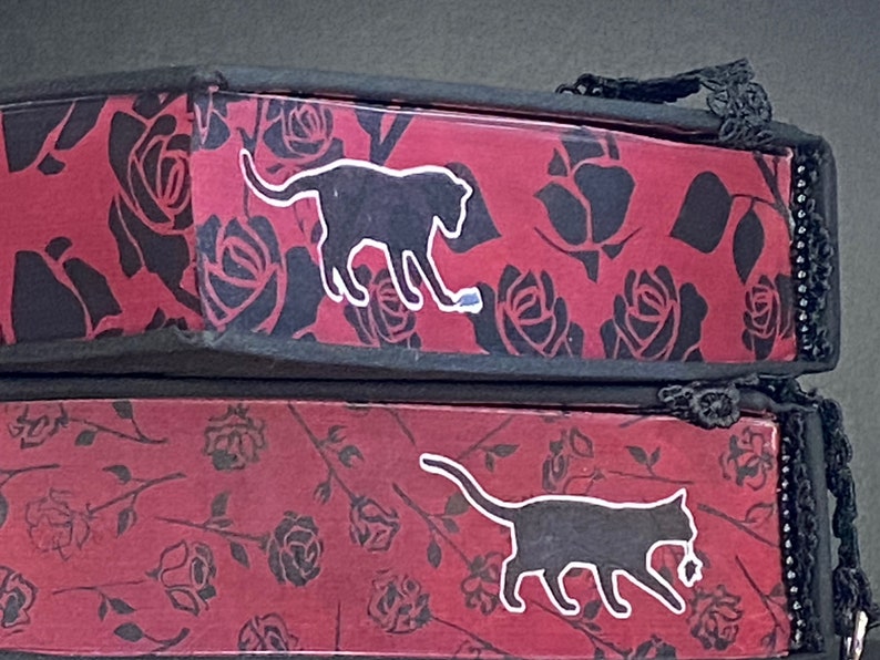 May include: Two stacked rectangular boxes featuring a red background with a black rose pattern. Each box displays a black cat silhouette with a white outline. The top box shows a cat playing with a small object, while the bottom box shows a cat walking.