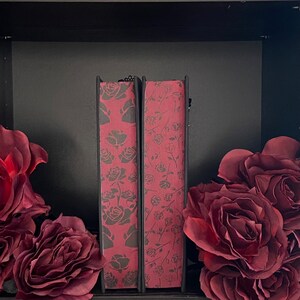May include: Two books with spines featuring a dark red background and a repeating pattern of dark gray roses. The books are positioned upright between two large, dark red artificial rose blossoms, creating a symmetrical floral arrangement.