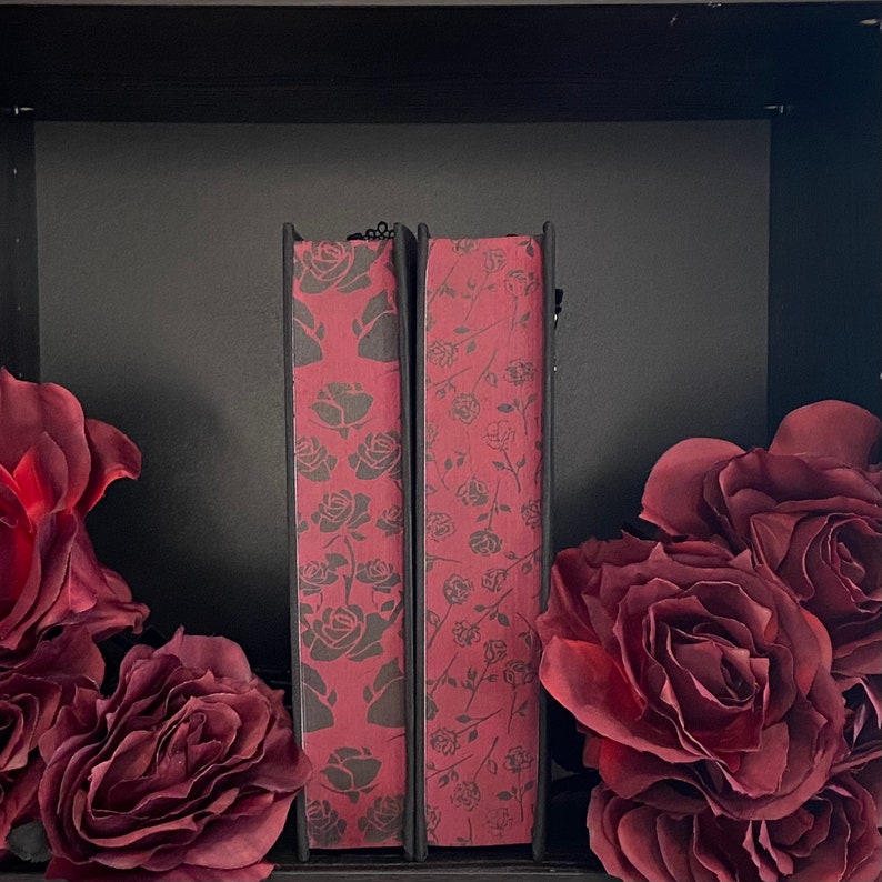 May include: Two books with spines featuring a dark red background and a repeating pattern of dark gray roses. The books are positioned upright between two large, dark red artificial rose blossoms, creating a symmetrical floral arrangement.