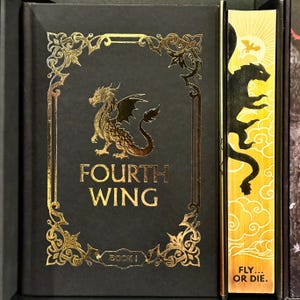 May include: A black hardcover book with a gold dragon design and the title "FOURTH WING" in gold lettering. The book is part of a boxed set and has a gold spine with the text "BOOK I". The book is next to a yellow bookmark with a gold dragon silhouette and the text "FLY... OR DIE."