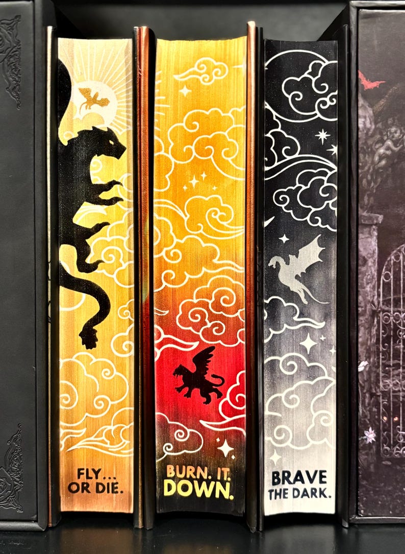 May include: Three fantasy novel book spines with a yellow, orange, and black background. The spines feature a dragon, a griffin, and a phoenix with the titles "Fly... or Die.", "Burn. It. Down.", and "Brave the Dark."