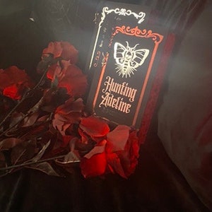 May include: A black book titled Hunting Adeline with a white moth illustration on the cover, framed by an orange border and ornate white scrollwork, positioned next to a cluster of deep red artificial roses with dark green stems and leaves.