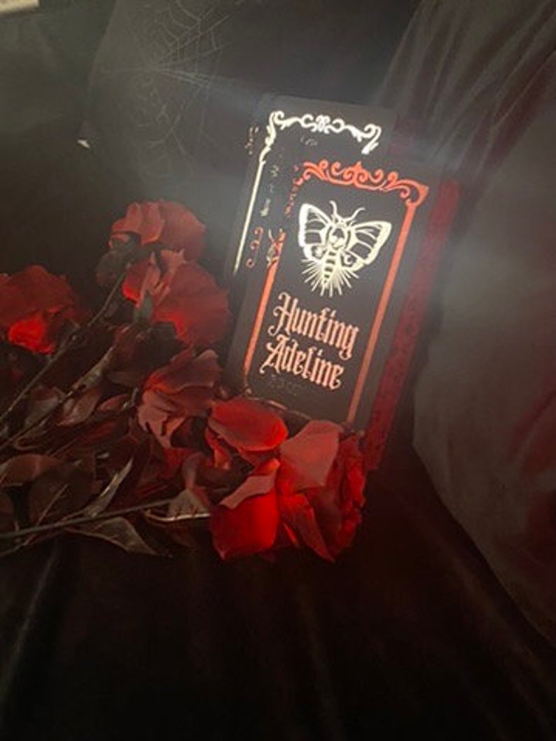 May include: A black book titled Hunting Adeline with a white moth illustration on the cover, framed by an orange border and ornate white scrollwork, positioned next to a cluster of deep red artificial roses with dark green stems and leaves.
