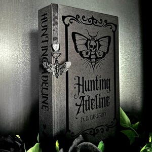 May include: A dark gray hardcover book titled Hunting Adeline by H.D. Carlton, featuring a moth illustration with a skull on the cover. A silver-toned metal moth charm hangs from a gray ribbon bookmark attached to the spine of the book.