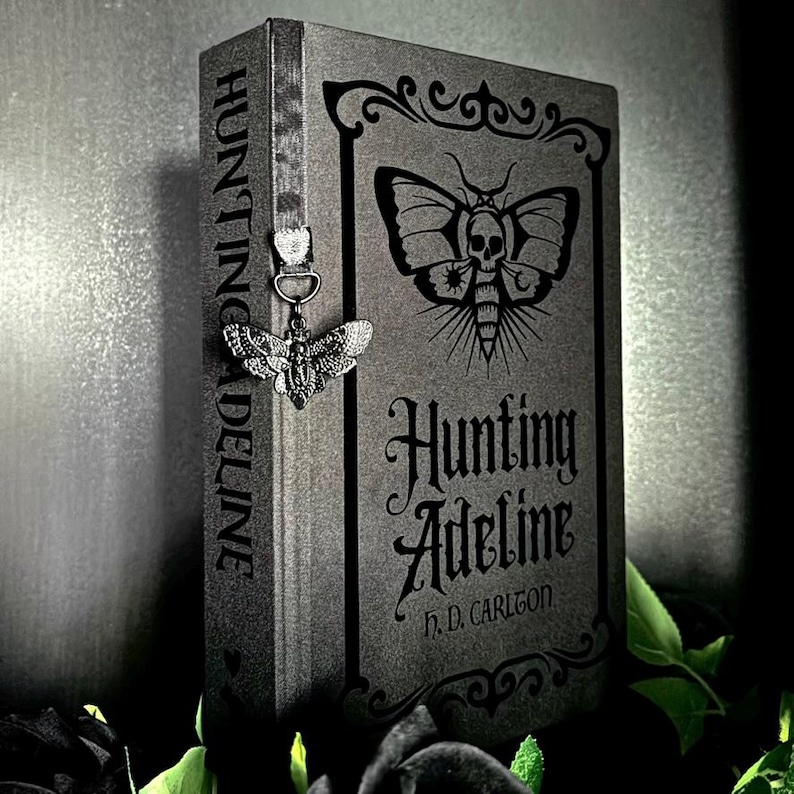 May include: A dark gray hardcover book titled Hunting Adeline by H.D. Carlton, featuring a moth illustration with a skull on the cover. A silver-toned metal moth charm hangs from a gray ribbon bookmark attached to the spine of the book.