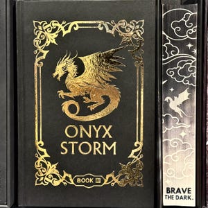 May include: A black book with a gold embossed cover featuring a dragon and the title "ONYX STORM BOOK III" with the words "BRAVE THE DARK." on the spine.