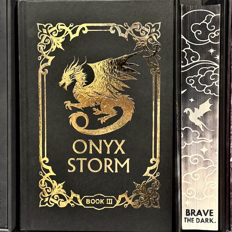 May include: A black book with a gold embossed cover featuring a dragon and the title "ONYX STORM BOOK III" with the words "BRAVE THE DARK." on the spine.