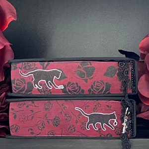 May include: Two stacked books with dark red covers featuring a black rose pattern. Each book spine displays a white silhouette of a cat. A black lace trim is attached to the right side of the spines, with a small silver cat charm hanging from it.