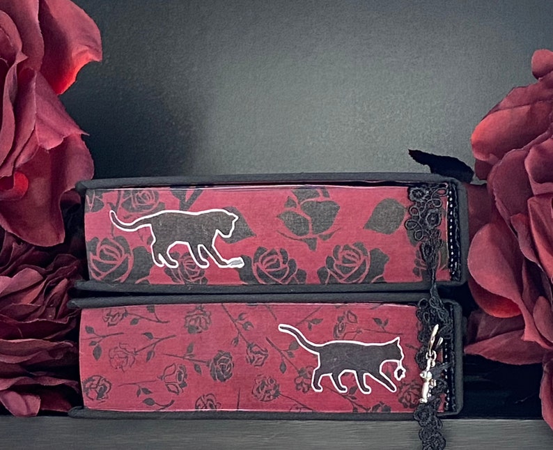 May include: Two stacked books with dark red covers featuring a black rose pattern. Each book spine displays a white silhouette of a cat. A black lace trim is attached to the right side of the spines, with a small silver cat charm hanging from it.
