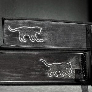 May include: Two stacked black books with white line art illustrations of cats on the spines. Each book features a distinct cat silhouette, one appearing to play with a small object, and both have black fabric binding and a black ribbon bookmark.