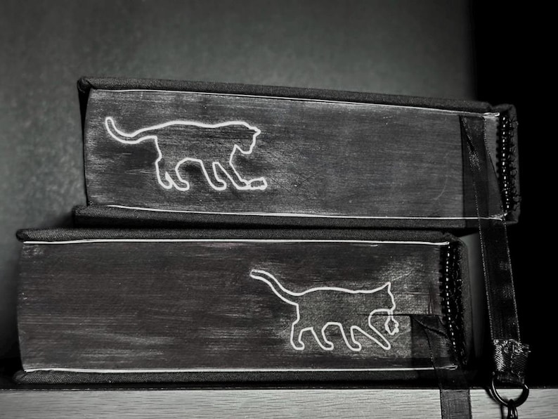 May include: Two stacked black books with white line art illustrations of cats on the spines. Each book features a distinct cat silhouette, one appearing to play with a small object, and both have black fabric binding and a black ribbon bookmark.