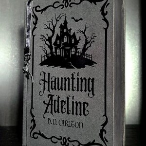 May include: A gray book titled Haunting Adeline by H. D. Carlton with a black graphic of a haunted house, bare trees, and a bat. The cover features a decorative black border and a thin gray ribbon bookmark with a small metal charm attached to the side.