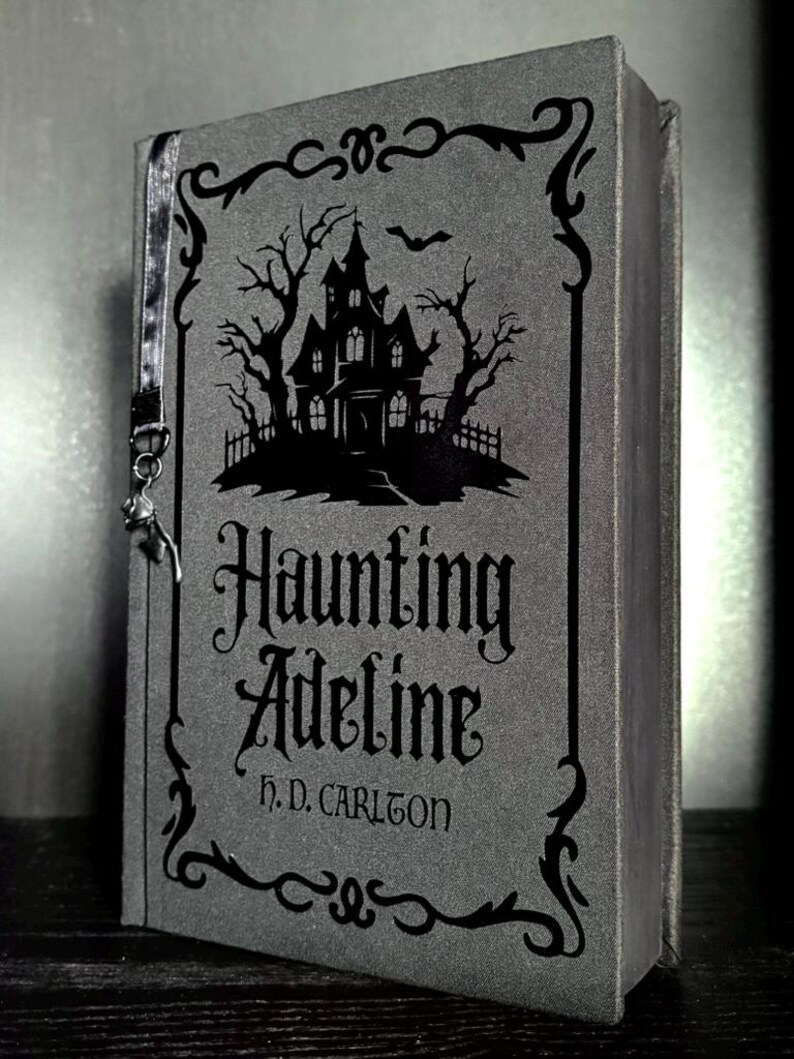 May include: A gray book titled Haunting Adeline by H. D. Carlton with a black graphic of a haunted house, bare trees, and a bat. The cover features a decorative black border and a thin gray ribbon bookmark with a small metal charm attached to the side.