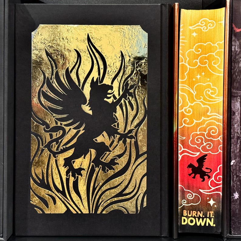 May include: A black and gold book cover with a black griffin silhouette in a gold flame design. The spine of the book has a gold and orange gradient with a black griffin silhouette and the text "BURN. IT. DOWN."