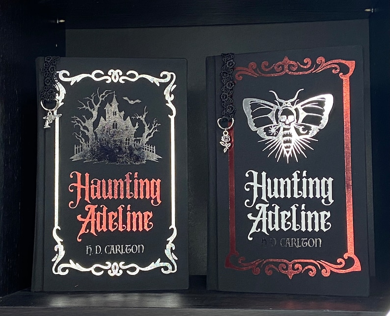 May include: Two black hardcover books titled Haunting Adeline and Hunting Adeline by H.D. Carlton. The first features a silver haunted house illustration, the second a silver moth with a skull. Both have black lace bookmarks with metal charms.