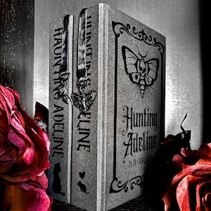 May include: A silver-toned book titled Hunting Adeline by H.D. Carlton with a moth and skull graphic on the cover. Two fabric bookmarks with metal charms, one shaped like a moth and one like a key, hang from the top edge of the book spine.