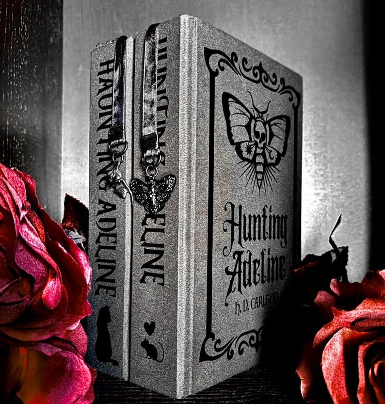 May include: A silver-toned book titled Hunting Adeline by H.D. Carlton with a moth and skull graphic on the cover. Two fabric bookmarks with metal charms, one shaped like a moth and one like a key, hang from the top edge of the book spine.