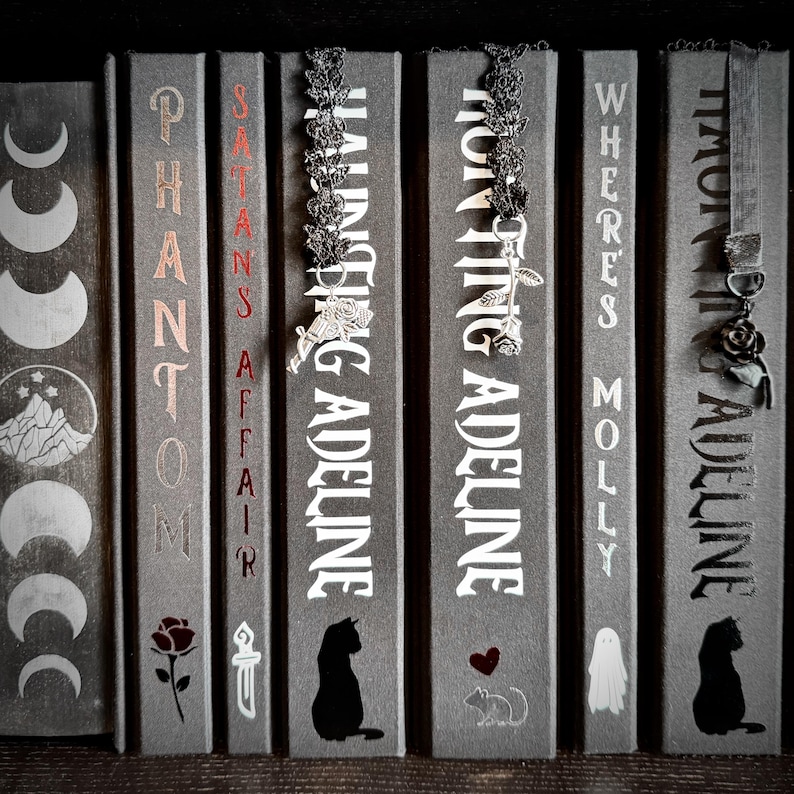 May include: A row of six gray books with white text and decorative elements including a moon cycle, a rose, a dagger, cats, a heart, a mouse, and a ghost. Three black lace bookmarks with silver charms hang from the tops of the book spines.