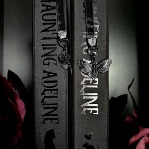 May include: Two black books titled Haunting Adeline with black ribbon bookmarks. One bookmark features a silver rose charm, and the other features a silver moth charm with a skull pattern. The book spines display the title and small animal silhouettes.