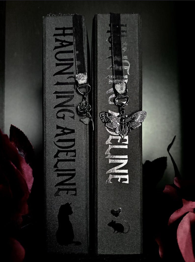 May include: Two black books titled Haunting Adeline with black ribbon bookmarks. One bookmark features a silver rose charm, and the other features a silver moth charm with a skull pattern. The book spines display the title and small animal silhouettes.