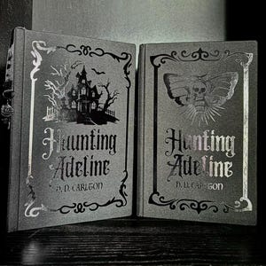 May include: Two dark gray hardcover books titled Haunting Adeline by H. D. Carlton. The left book features a silver foil illustration of a haunted house and trees. The right book displays a silver foil moth with a skull pattern on its wings.