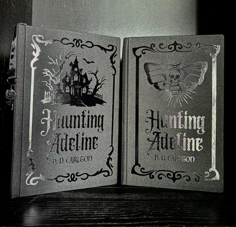 May include: Two dark gray hardcover books titled Haunting Adeline by H. D. Carlton. The left book features a silver foil illustration of a haunted house and trees. The right book displays a silver foil moth with a skull pattern on its wings.
