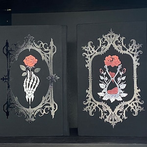 May include: Two black books with silver ornate frames on the covers. The left book features a white skeleton hand holding a red rose. The right book displays an hourglass with red sand, surrounded by red roses and silver leaves, all in a detailed style.