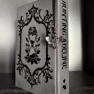 May include: A gray hardcover book titled Hunting Adeline with a black ornate frame design on the cover featuring a rose and an hourglass. A black ribbon bookmark hangs from the top, attached to a silver-toned moth charm with intricate wing details.