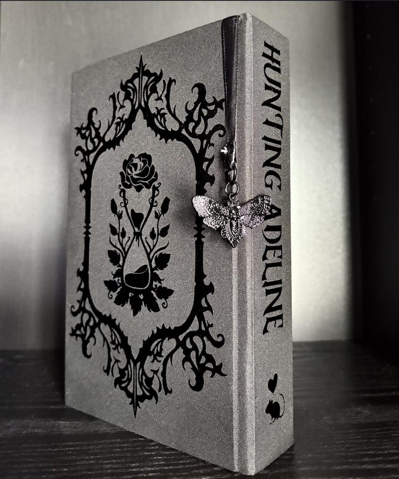 May include: A gray hardcover book titled Hunting Adeline with a black ornate frame design on the cover featuring a rose and an hourglass. A black ribbon bookmark hangs from the top, attached to a silver-toned moth charm with intricate wing details.