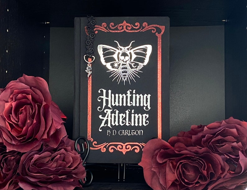 May include: A black hardcover book titled Hunting Adeline by H D Carlton with a silver moth and skull graphic on the cover. The book features a metallic red border and a black lace bookmark with a silver rose charm attached to the top edge.