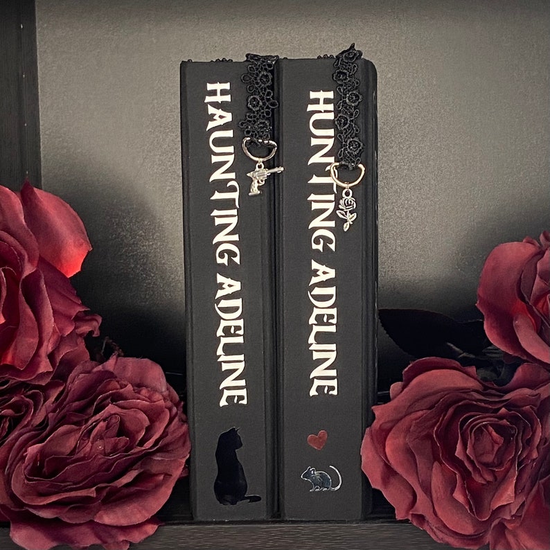 May include: Two black books titled Haunting Adeline with white text, each featuring a black lace bookmark. One bookmark has a silver pistol charm, the other a silver rose charm. The book spines display a black cat silhouette and a mouse with a heart.