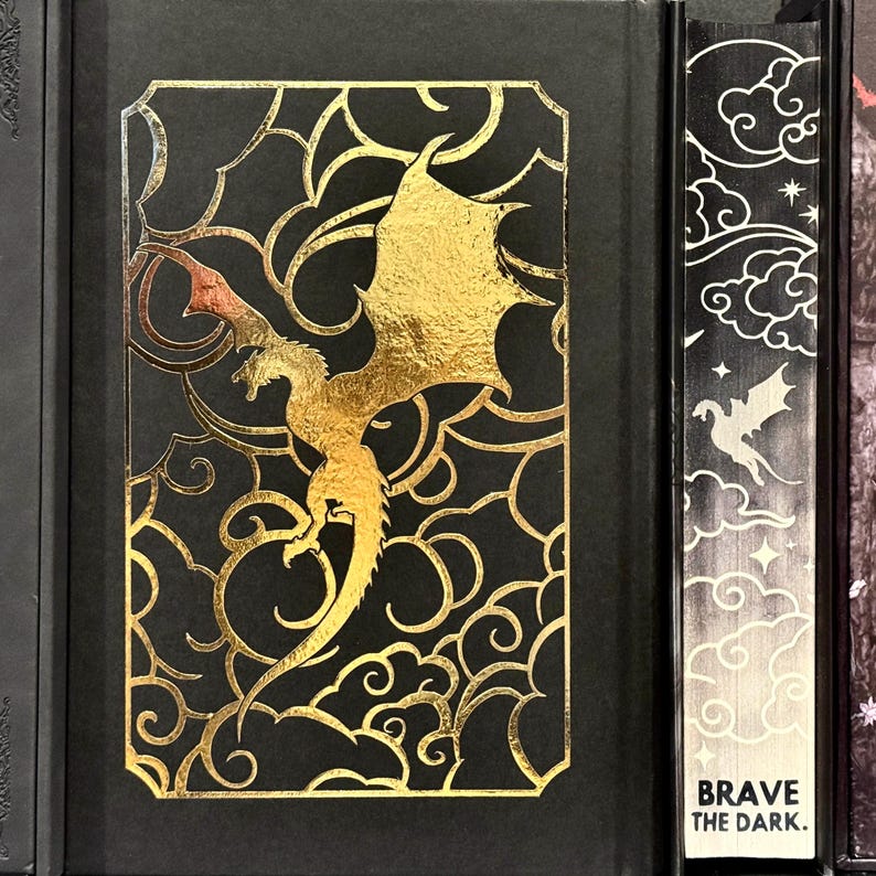 May include: A black hardcover book with a gold dragon design on the front cover. The dragon is flying against a background of gold clouds. The spine of the book has the text "BRAVE THE DARK." in white.