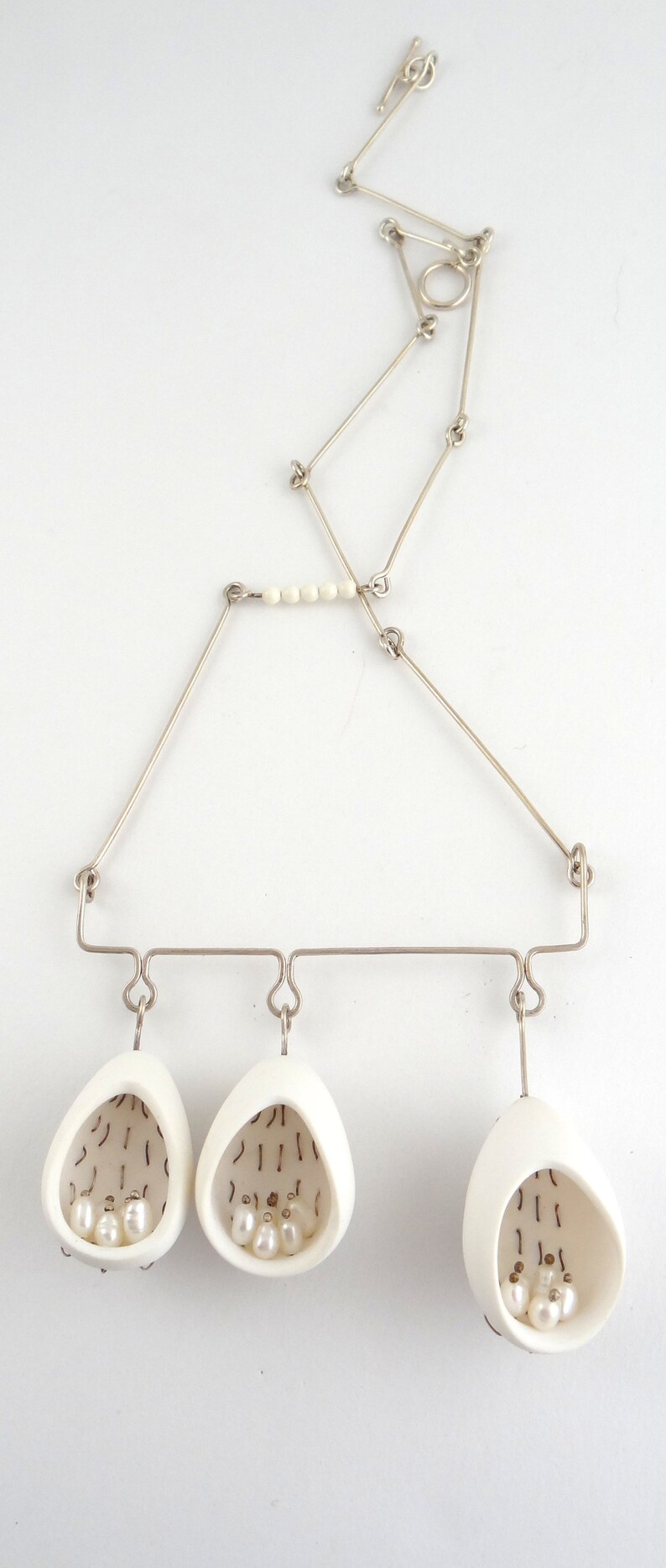 May include: A silver necklace with three white ceramic pendants, each containing small white pearls. The pendants are shaped like teardrops and have a delicate, intricate design.